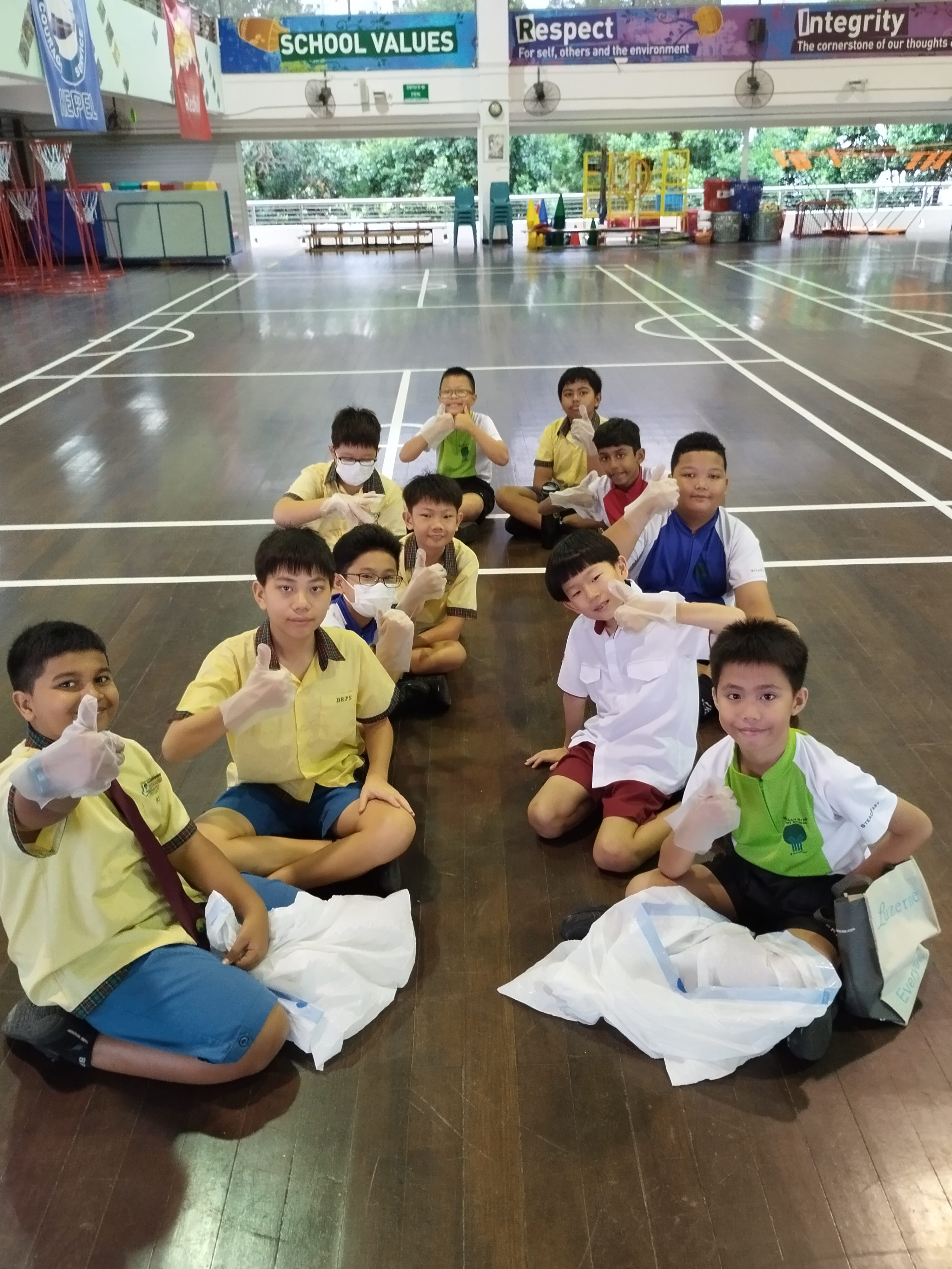 A group of school children sit on a gym floor wearing gloves and giving thumbs up, with bags in front of them.
